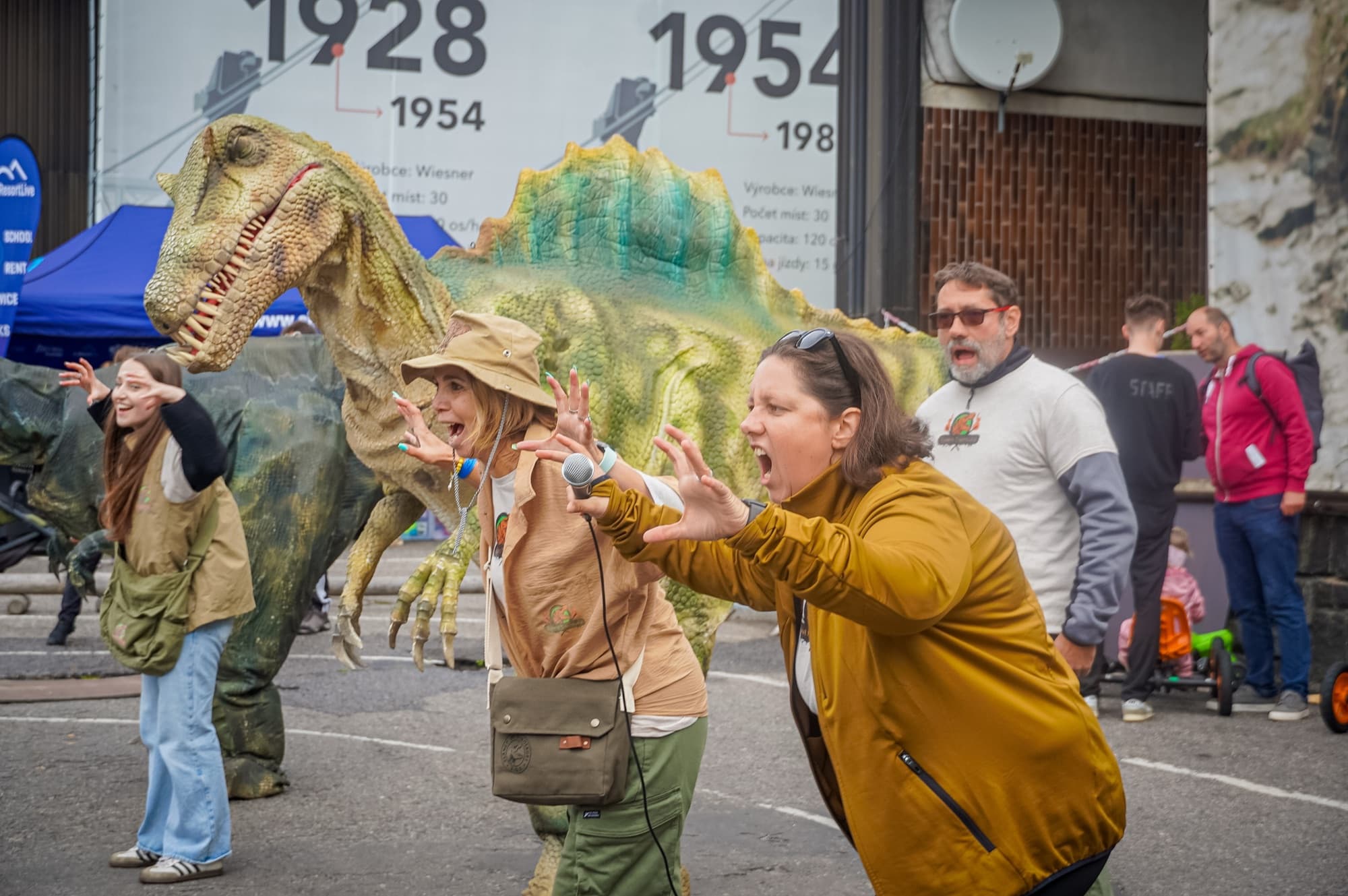 Animátorka s mikrofonem vede program, za ní stojí dva dinosauři