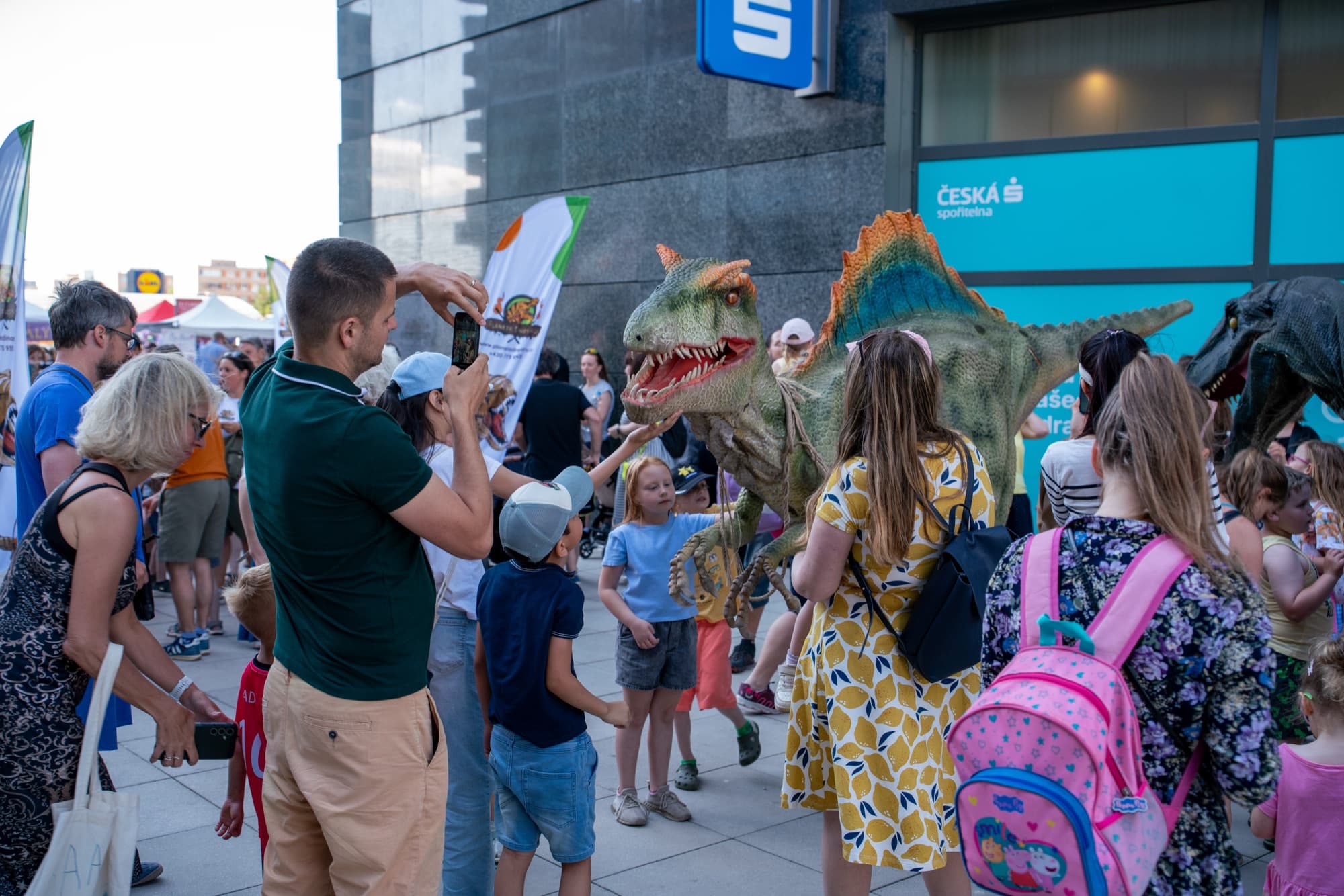 Dinosaurus prochází davem lidí na městské akci, rodiče ho fotí na telefon