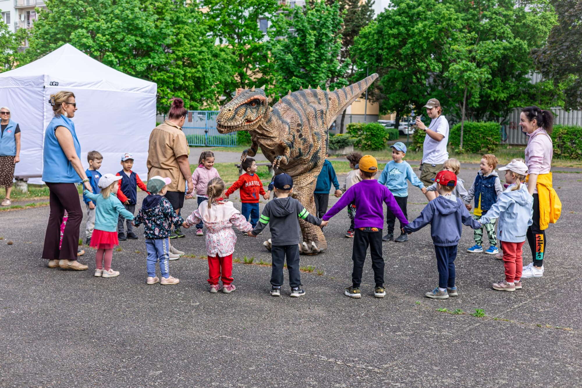 Děti z mateřské školy stojí v kruhu kolem dinosaura na hřišti
